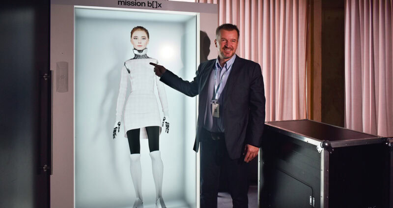 Filip Linek standing next to a case featuring BE-A, a humanoid AI Receptionist, showcasing the future of the hotel industry.