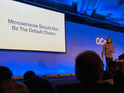A speaker stands on stage at QCon London 2020 next to a screen displaying the text Microservices Should Not Be The Default Choice.