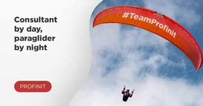 A consultant soars as a paraglider under a red #TeamProfinit wing, cutting through the cloudy sky.