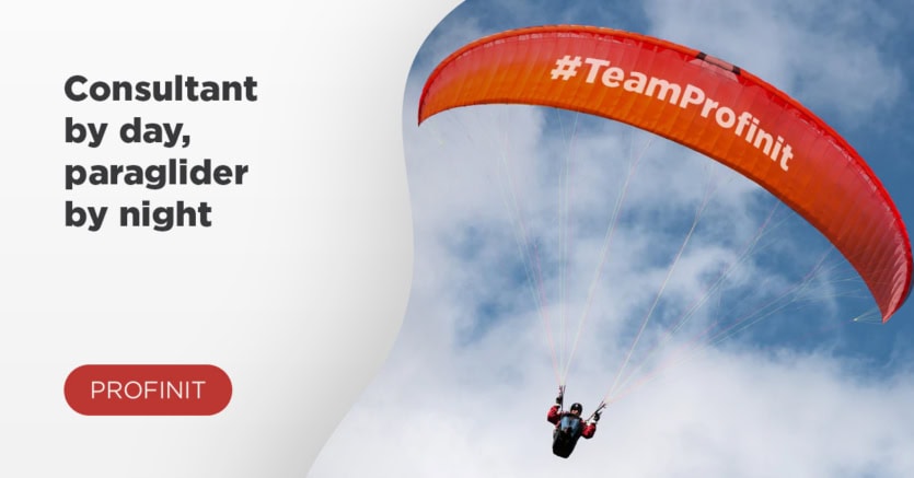 A consultant soars as a paraglider under a red #TeamProfinit wing, cutting through the cloudy sky.
