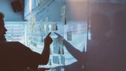 Two people discuss and point at sticky notes arranged on a whiteboard, collaborating on Agile tasks.