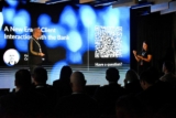 A presenter stands on stage beside a large screen displaying a talk titled A New Era: Client Interaction with the Bank.