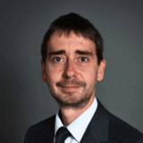 A man with short brown hair, light skin, and a trimmed beard wears a gray suit, white shirt, and dark tie, posing confidently against a plain dark background—reflecting the professionalism often seen in systems modernization leaders.